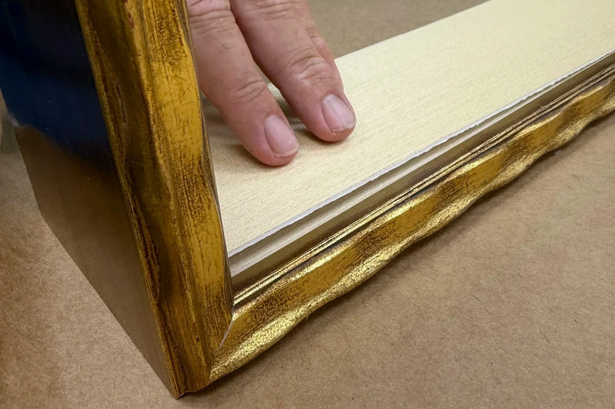 Close-up of a person's hand with fingers touching the surface of a gold-colored picture frame. The frame appears vintage with intricate detailing, and the background shows a beige surface.