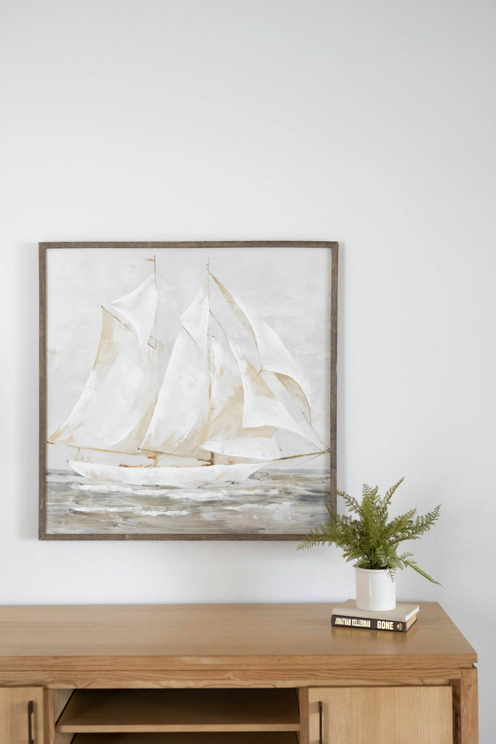 A framed painting of a white sailboat with white sails on water in a minimalist apartment setting. The painting is hung on a white wall above a wooden sideboard. To the right of the painting is a small potted fern. On the sideboard are two books, one with the title "GONE" by Jonathan Kellerman and a white ceramic pot with a green fern.