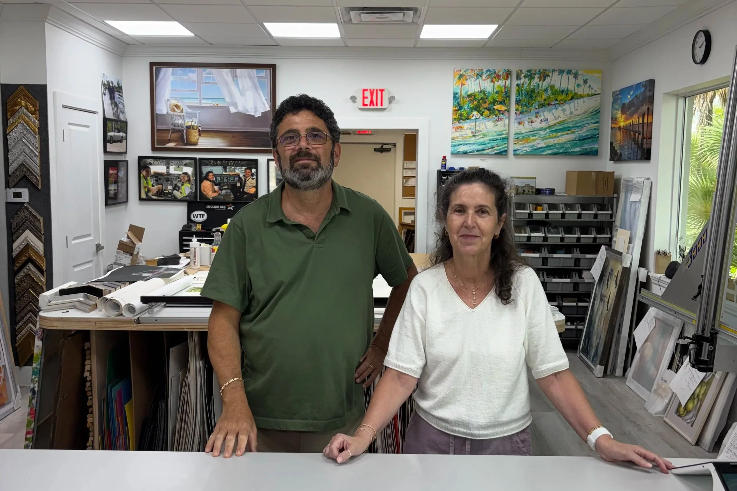 Paul and Valerie Roshberg standing side by side in an art supply store, with framed artwork, art supplies, and samples on display behind them.