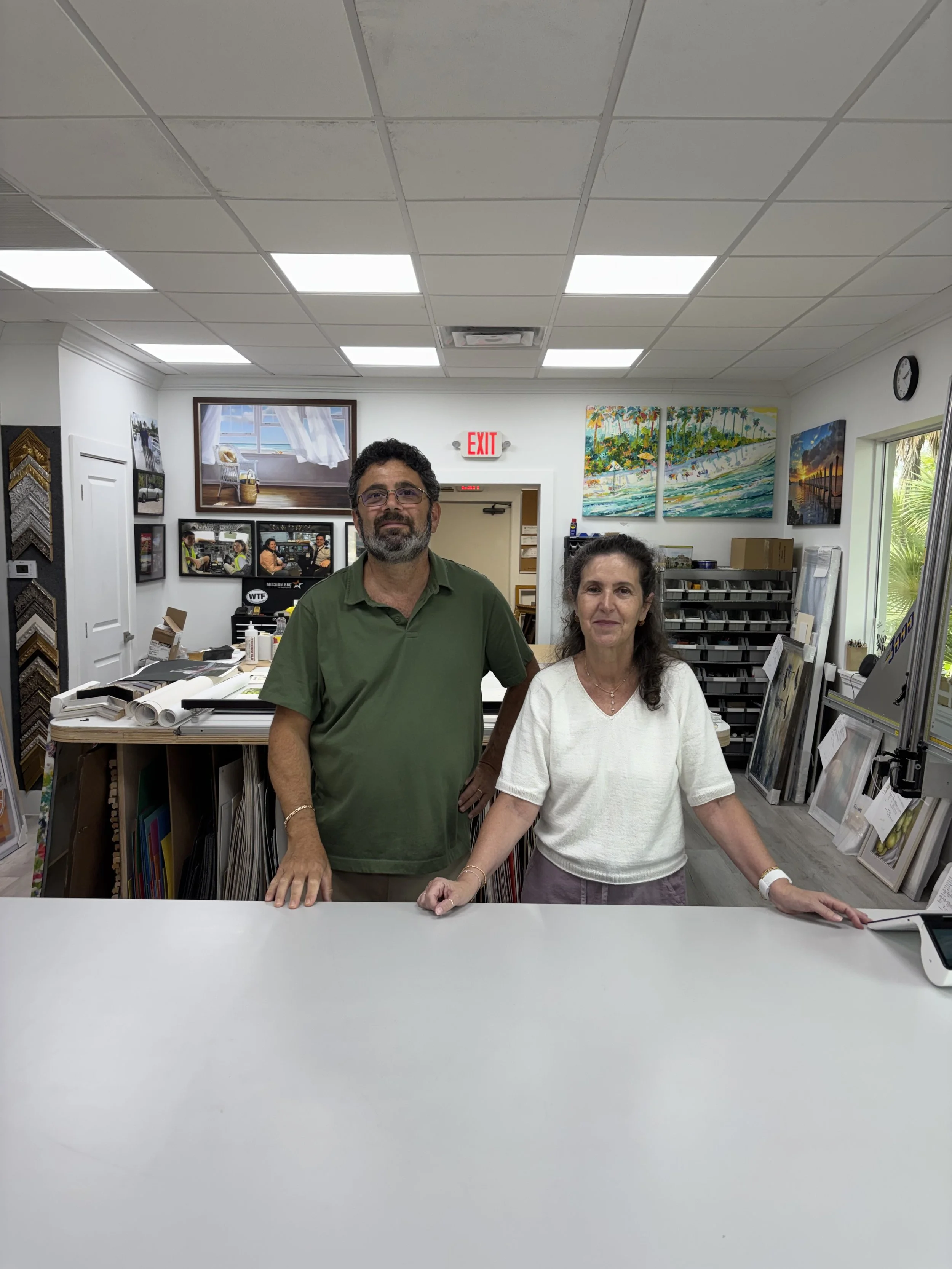 A man and a woman standing at a white counter in an art store, surrounded by paintings and framing supplies, with a window and paintings hanging on the wall behind them.