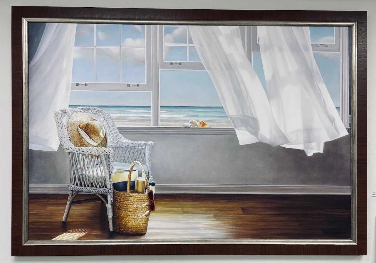 A painting of a seaside scene viewed through large open windows with white curtains. There is a white wicker chair with a straw hat and sunglasses on it, and a wicker basket on the floor next to it filled with beach items.