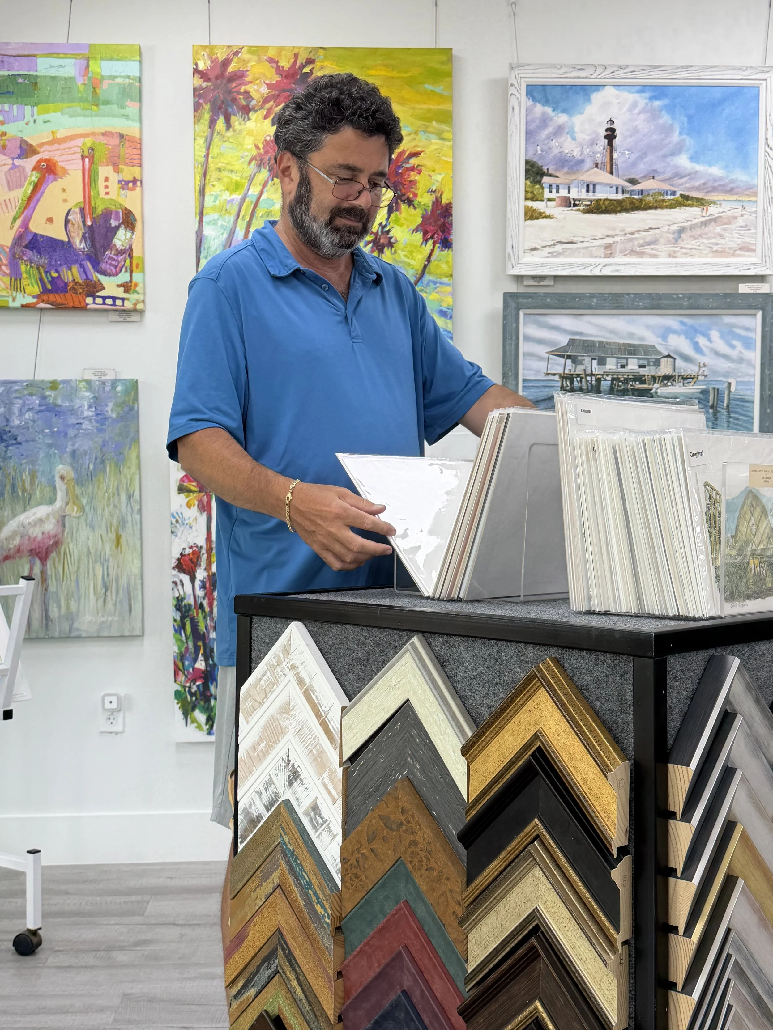 A man browsing through picture framing samples at an art gallery or framing shop, with colorful paintings hanging on the wall behind him.