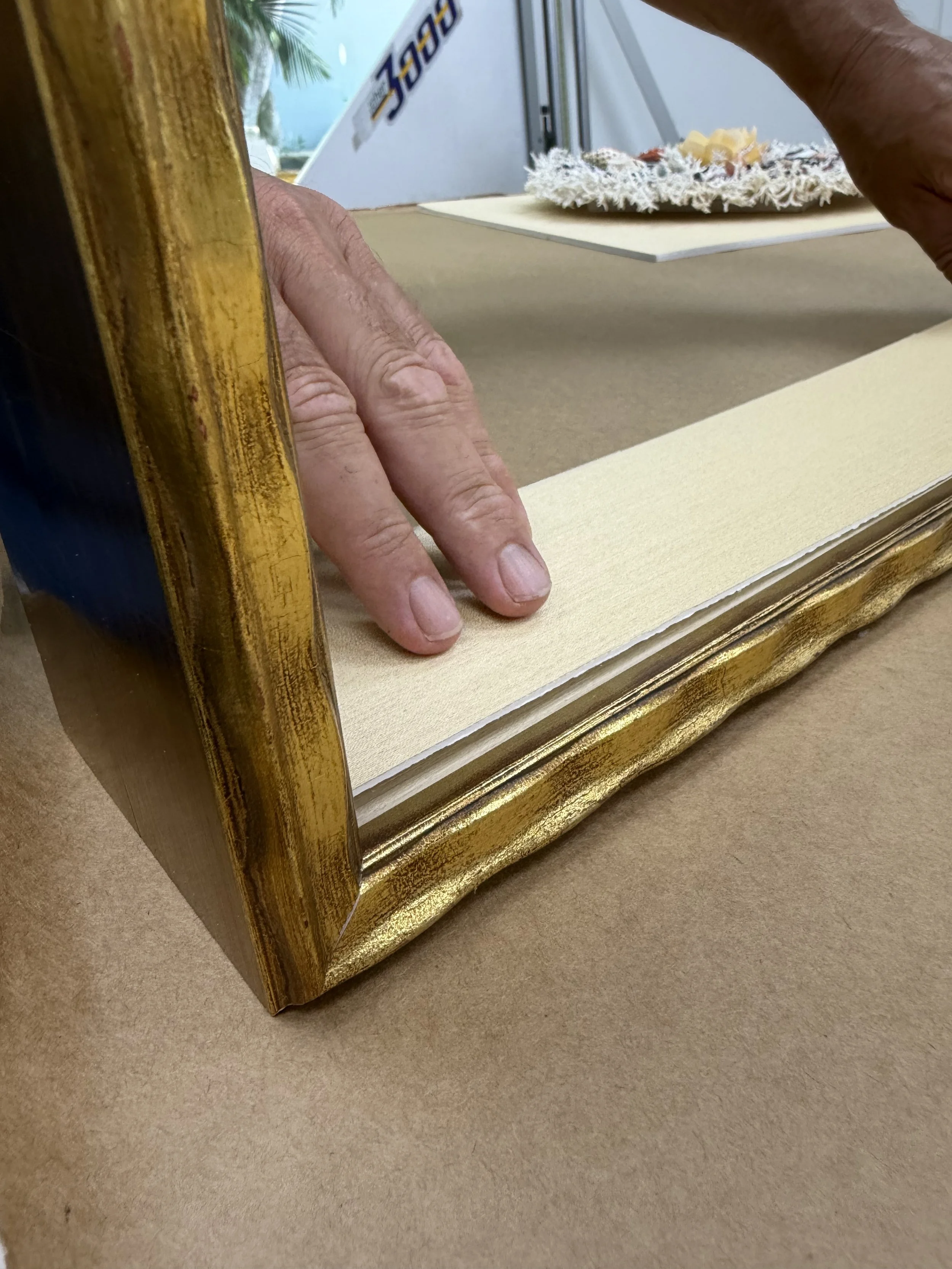 Close-up of a person's hand with visible wrinkles and grayish skin, adjusting or setting a gold-colored picture frame on a beige surface.