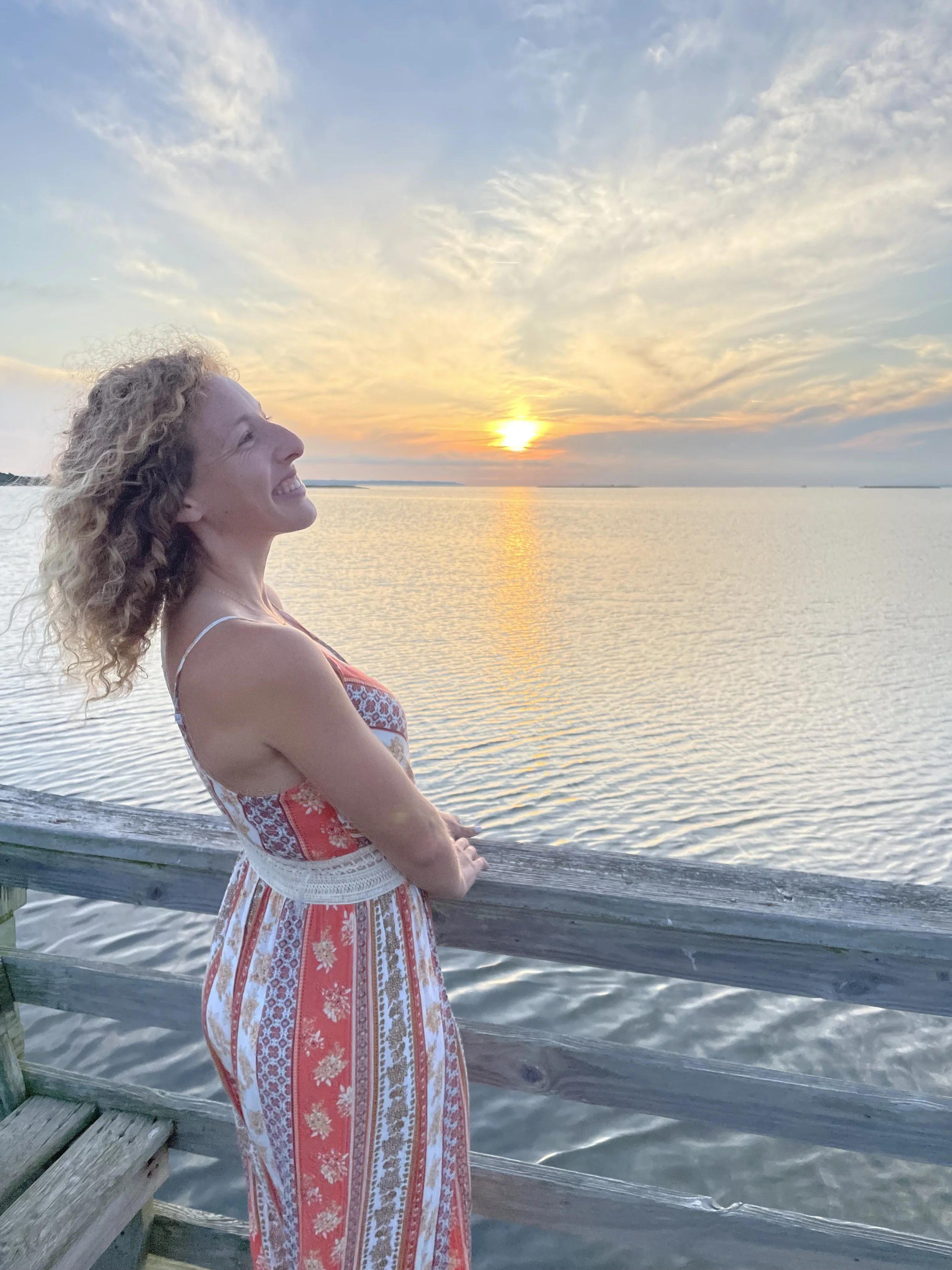 A woman with curly blonde hair smiling as she looks toward the sunset over the water, standing by a wooden railing on a pier during the evening.