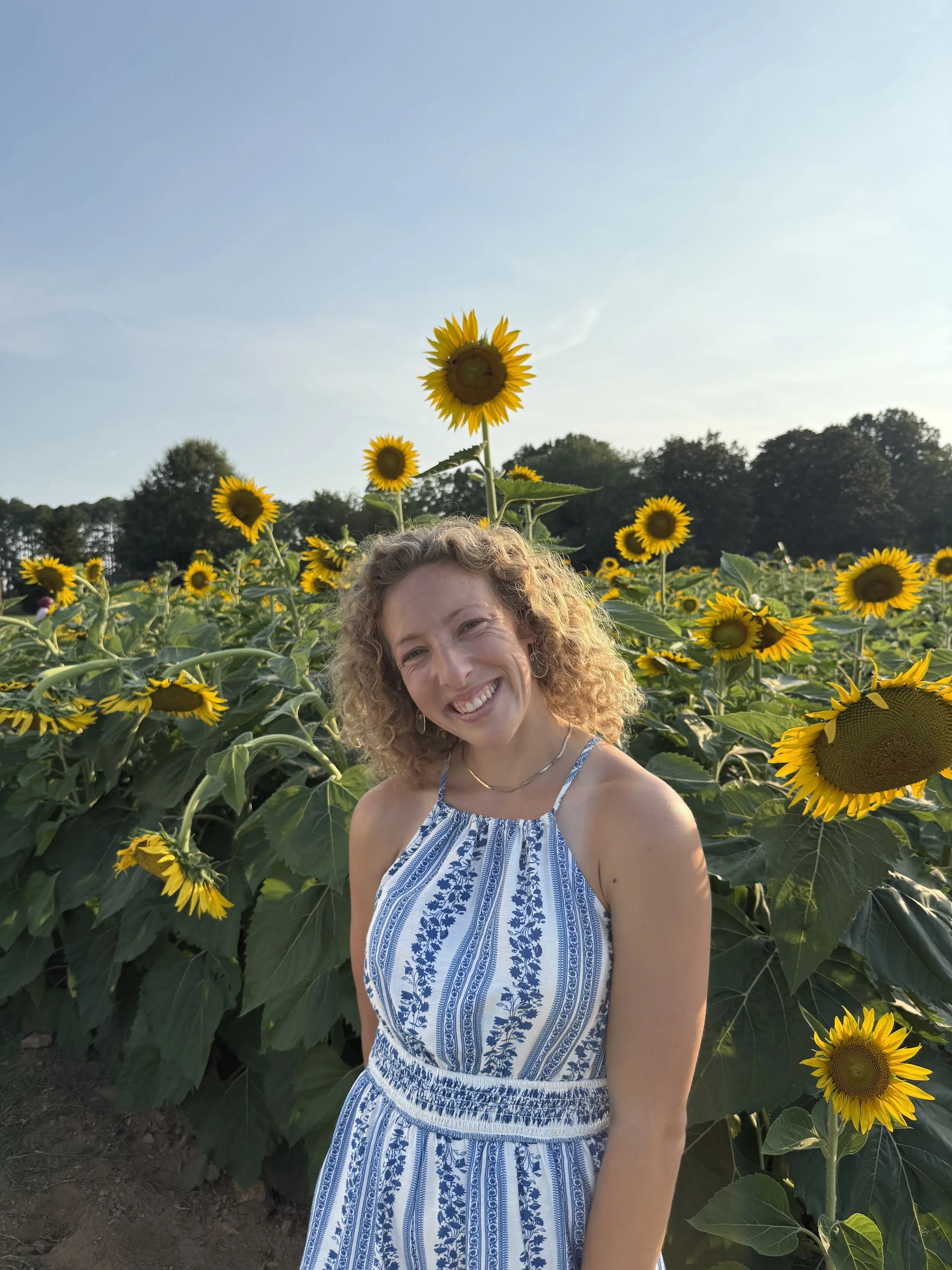 A woman with curly hair smiling in a sunflower field during daytime, wearing a blue and white sleeveless dress