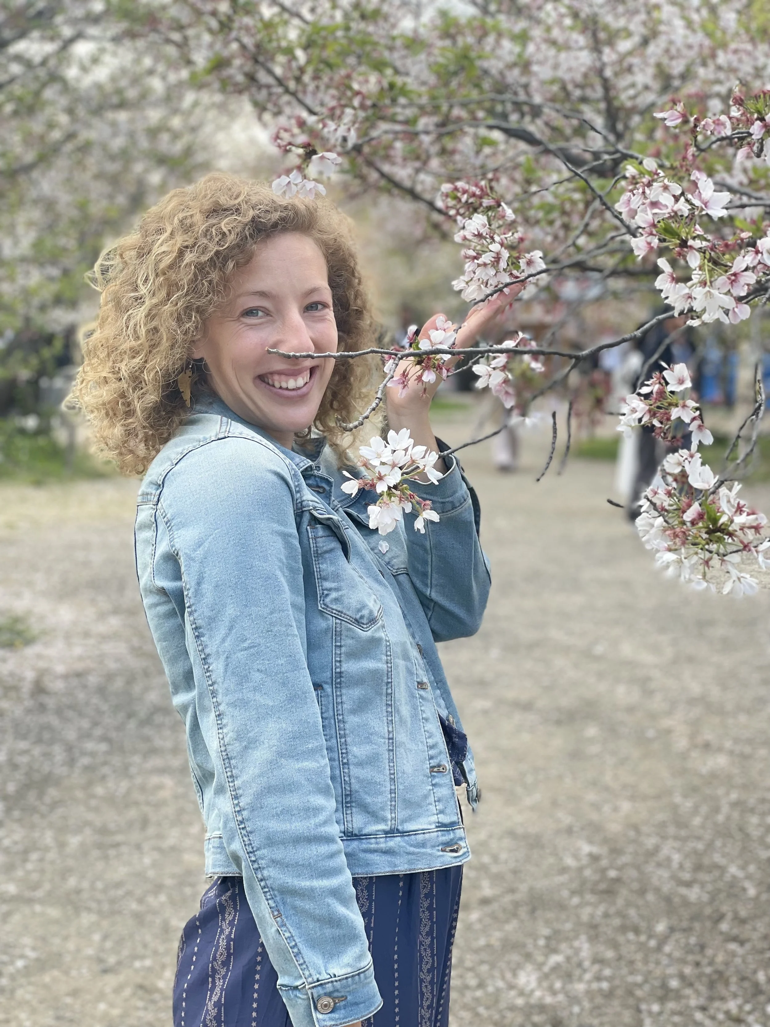 A woman with curly blonde hair smiling and holding a branch of pink and white cherry blossoms in a park.