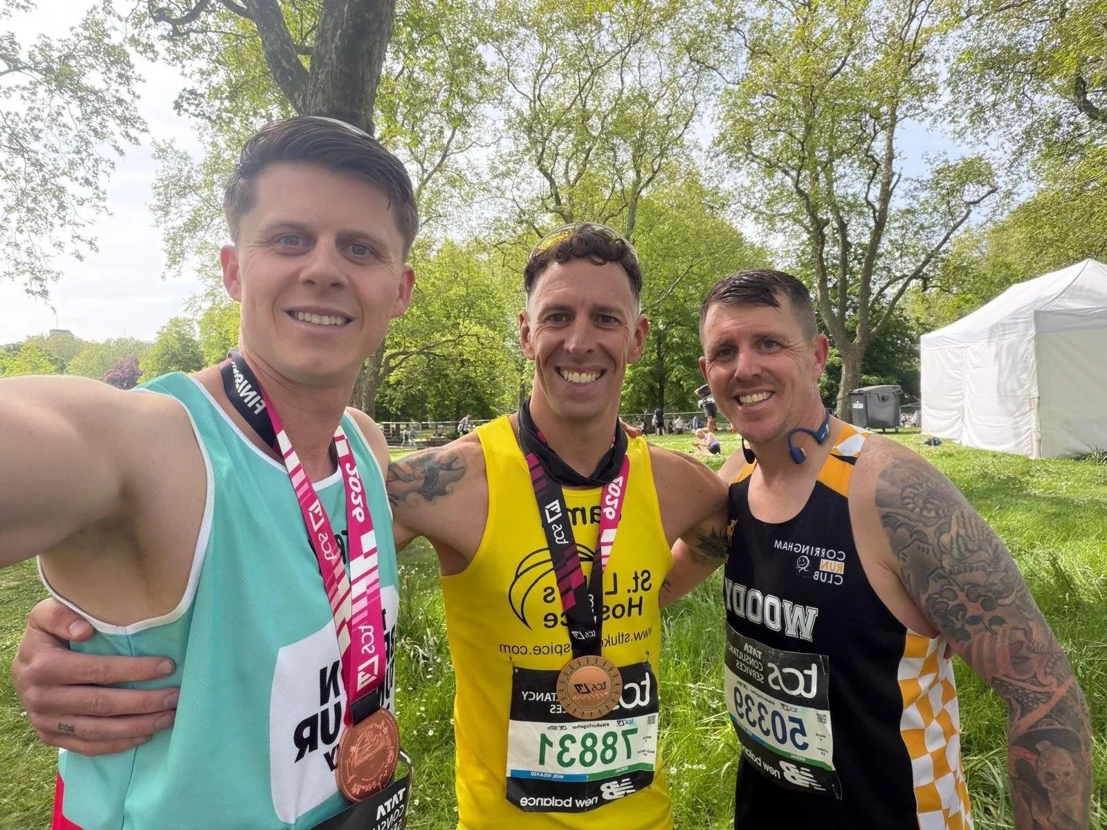 Three male runners posing for a photo after the London Marathon, wearing medals, with trees and a white tent in the background.