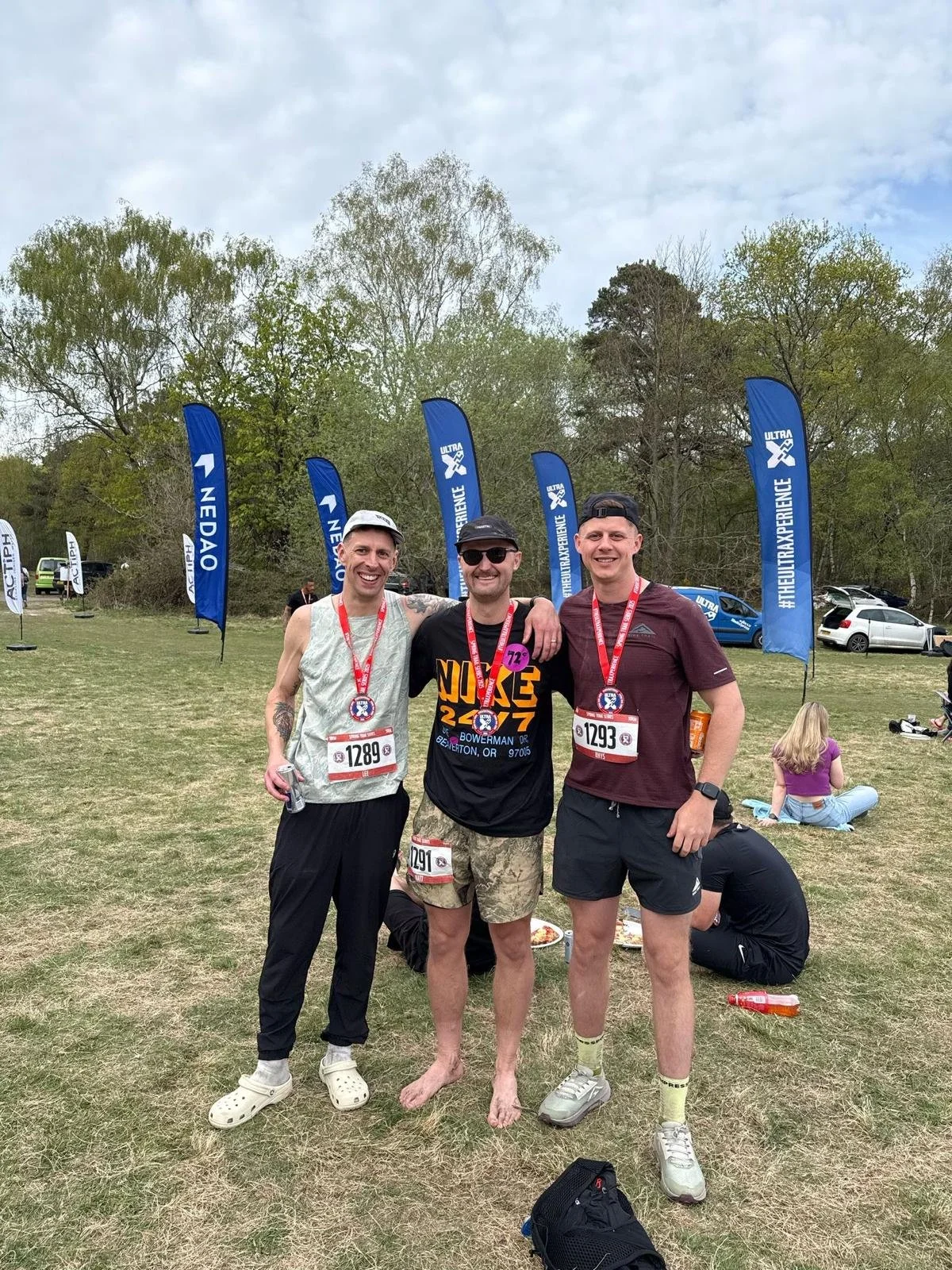 Three smiling men standing together outdoors after a race, wearing race bibs and medals. They are surrounded by event flags and parked cars, with a woman and a person sitting on the grass in the background.