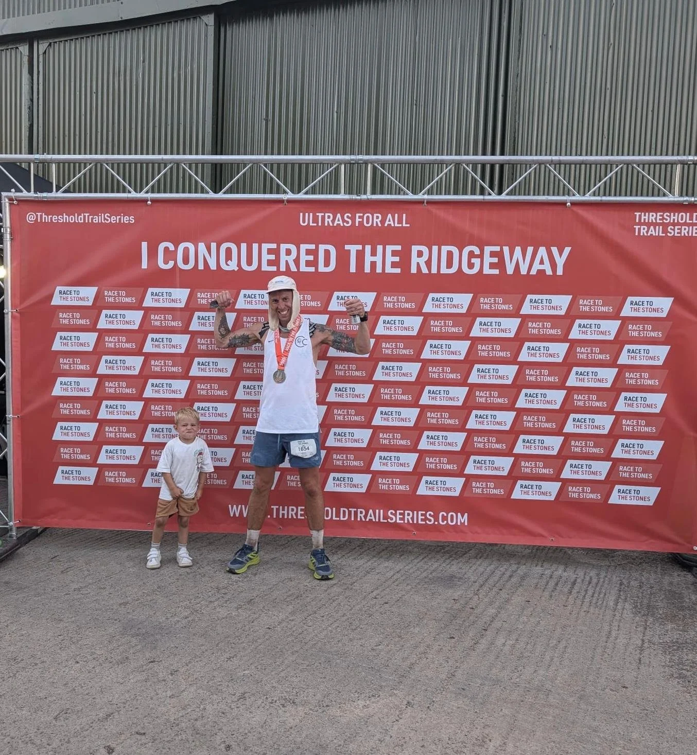 A man and a young boy standing in front of a red banner at a trail running event. The man is wearing a white cap, a white tank top, shorts, and running shoes, and has a medal around his neck. The boy is wearing a white t-shirt, beige shorts, and sneakers. The banner displays the phrase "I Conquered the Ridgeway," with additional text and logos promoting the event.