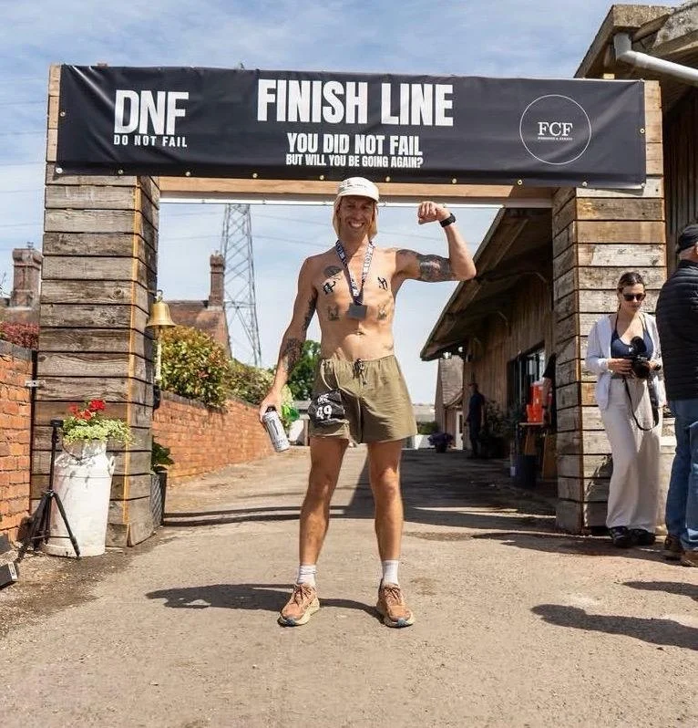 A man standing at the finish line of a race, smiling and flexing his arm. He is wearing a white cap, a medal around his neck, shorts, and running shoes. The finish line banner reads 'DNF Finish Line, You Did Not Fail, But Will You Be Going Again?'