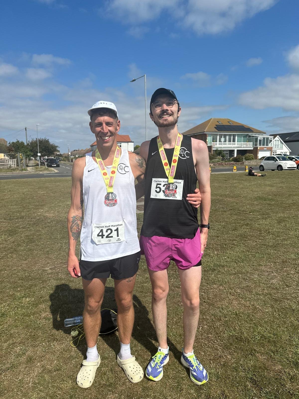 Two male runners standing outdoors after completing a half marathon, wearing medals and race bibs, smiling at the camera, with a residential neighborhood in the background.