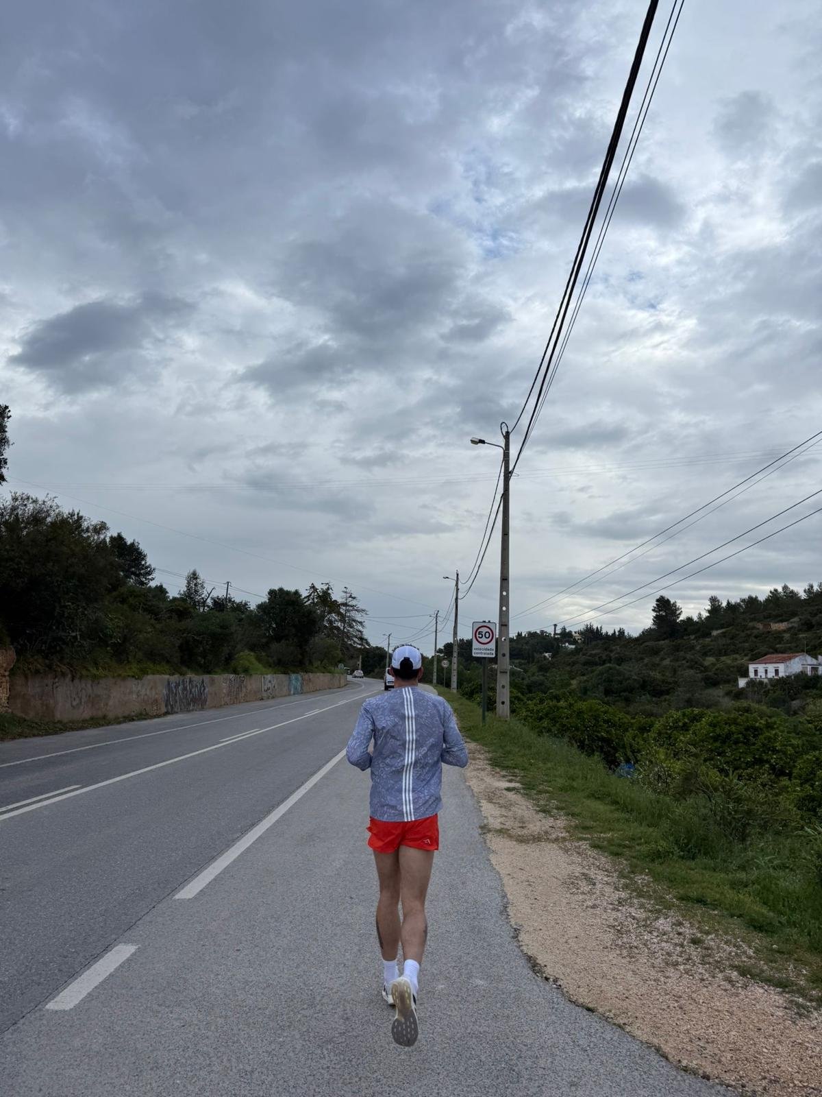 A person running on a rural road during a cloudy day, wearing a white cap, a light blue shirt, red shorts, and white shoes, with trees, grassy areas, and houses in the background.