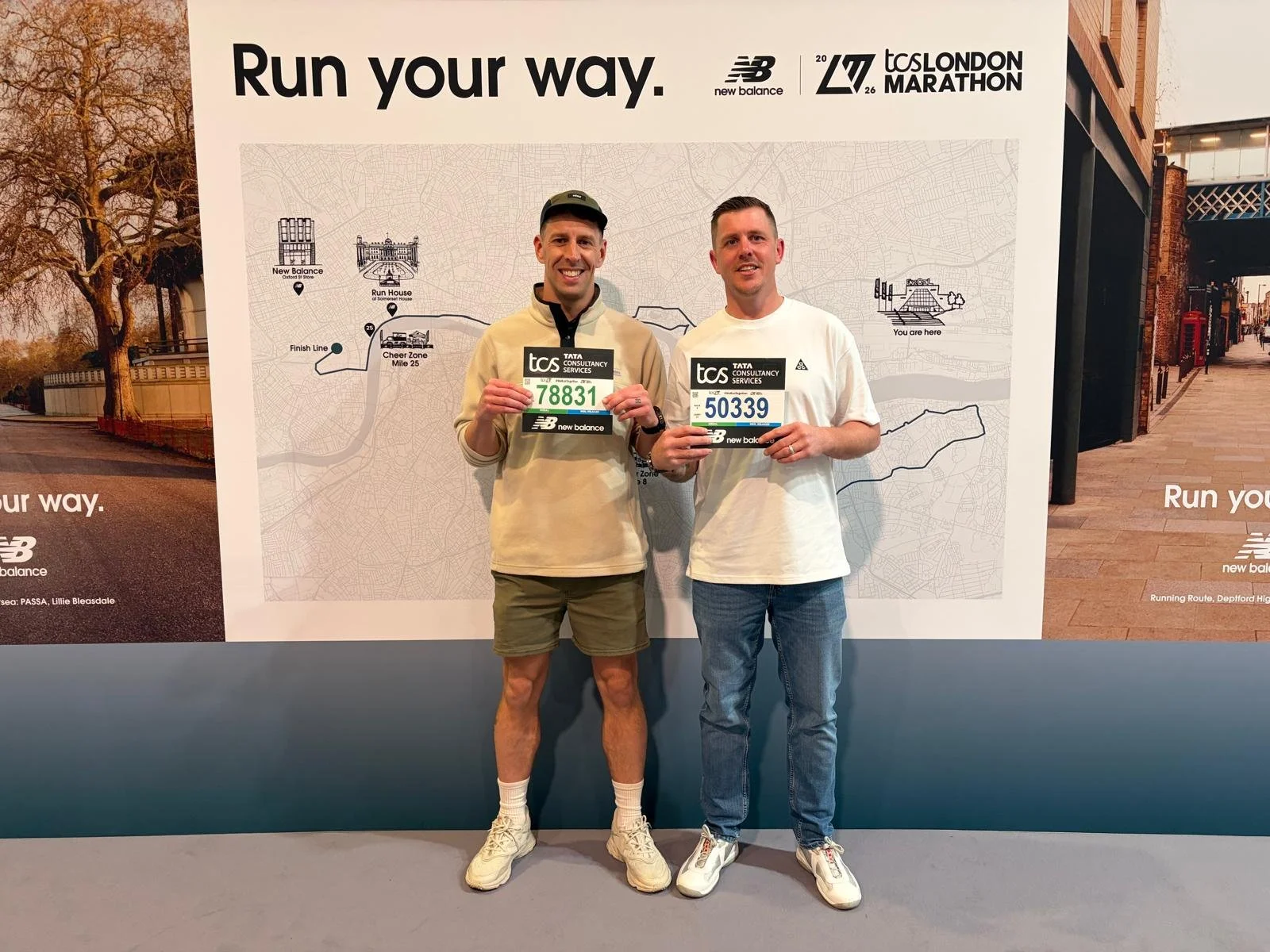 Two men holding marathon bibs in front of a poster at a marathon event. The poster reads 'Run your way' and has a map with route details for the London Marathon.