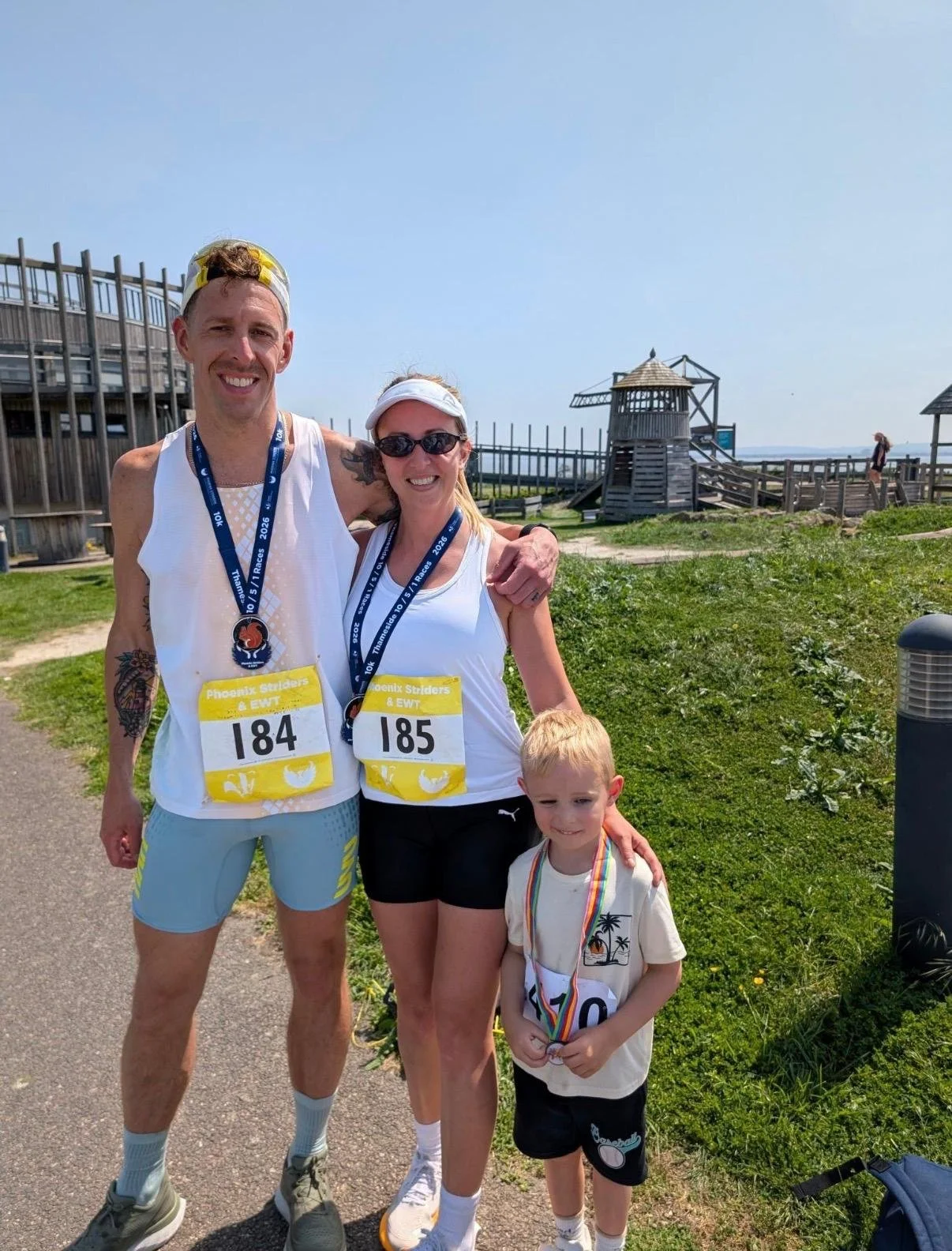 Two adults and a child standing outdoors after running a race, smiling at the camera. The adults are wearing race bibs numbered 184 and 185, with medals around their necks. The child has a medal and is holding a small object. There is a wooden structure and a grassy area in the background, with a blue sky overhead.