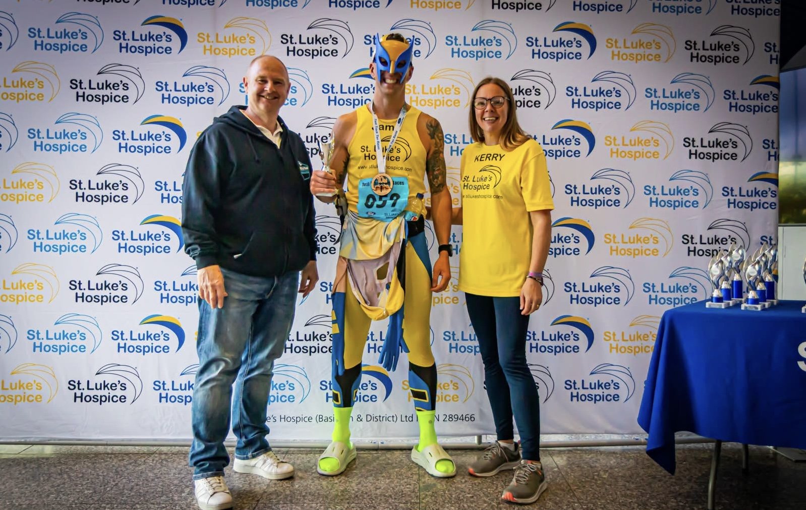 Three people standing in front of a St. Luke's Hospice backdrop, with a runner in the middle wearing a mask and holding a medal, celebrating their achievement at a charity event.