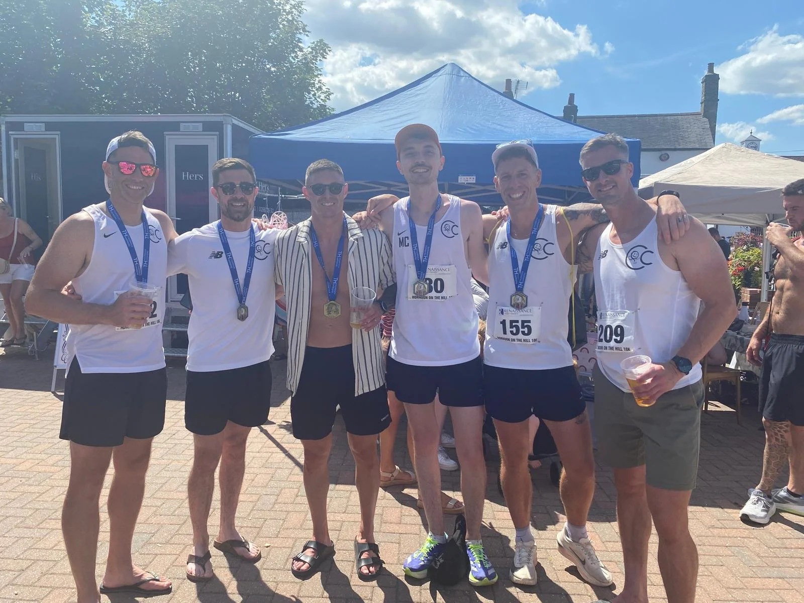 A group of six men in athletic gear celebrating after a race, wearing medals around their necks, standing outdoors in front of a blue tent on a sunny day. Some hold drinks, and some have race bibs pinned to their shirts.