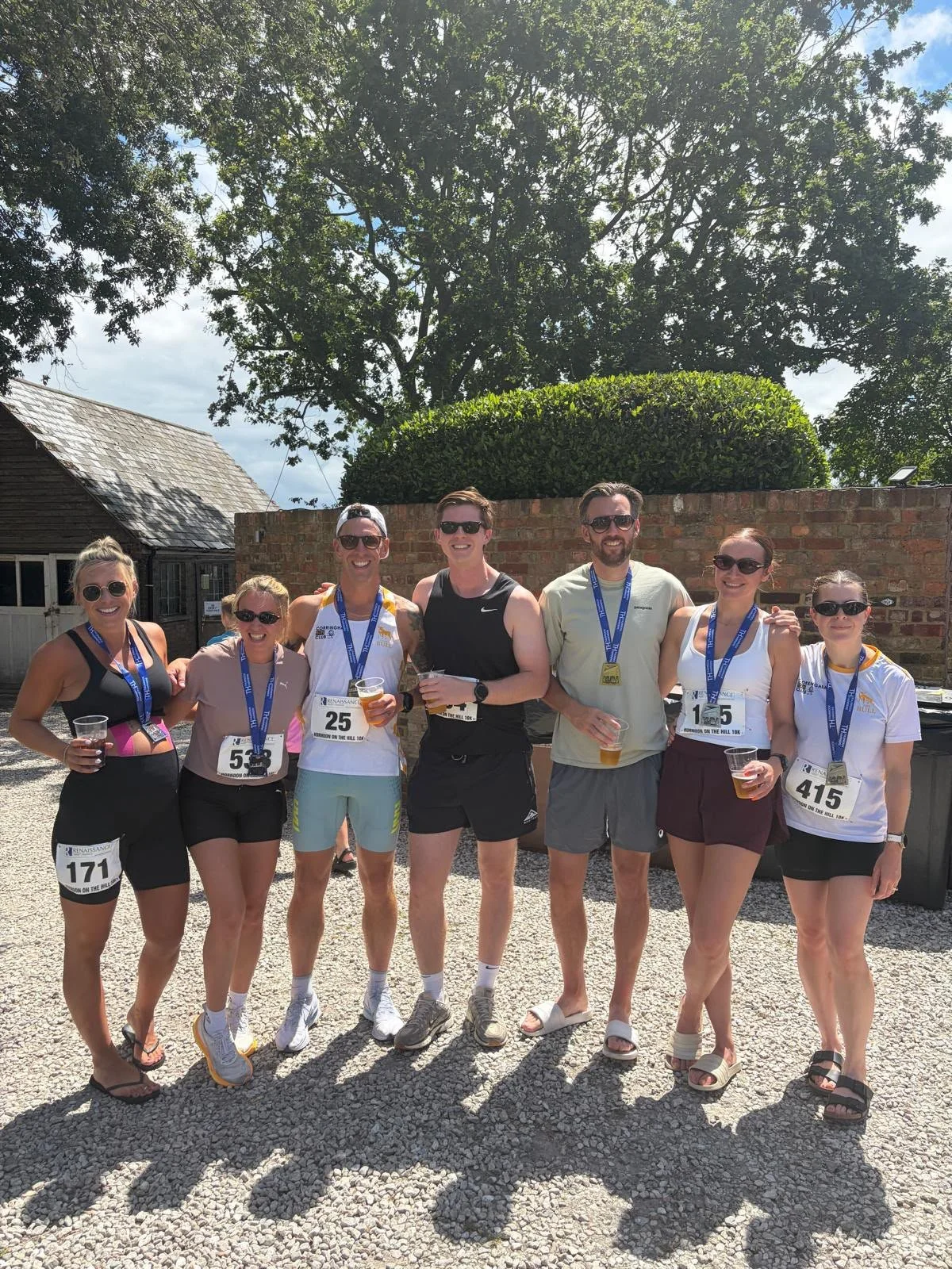 Group of seven smiling runners with medals and drinks, standing outdoors after a race, with trees, a brick wall, and a barn in the background.