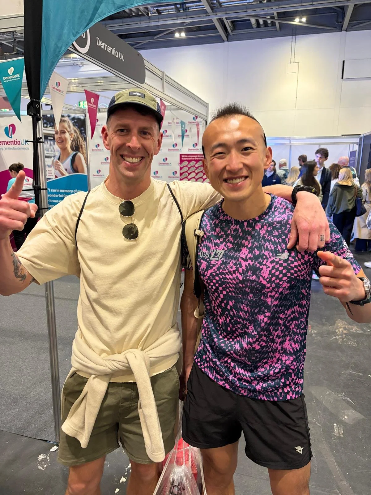 Two men smiling with arms around each other in a crowd at an indoor event. The man on the left is wearing a beige t-shirt, sunglasses hanging from his shirt, and a cap. The man on the right is wearing a patterned pink and black t-shirt and black shorts.