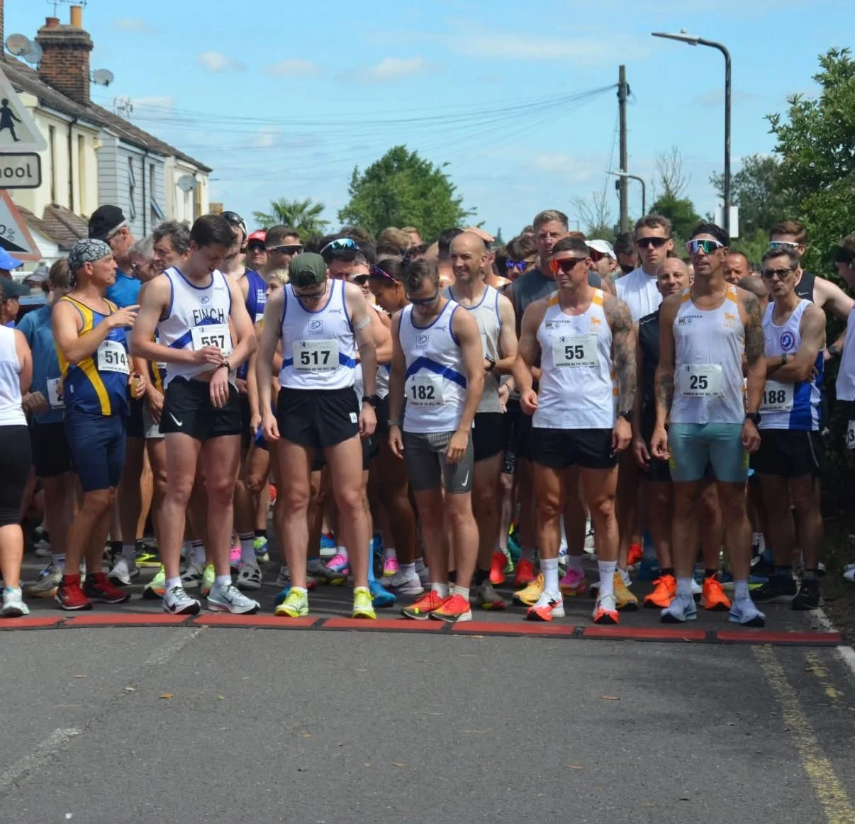 A group of runners gathered at the starting line for a race, wearing athletic gear and race bibs, in a suburban neighborhood on a clear day.