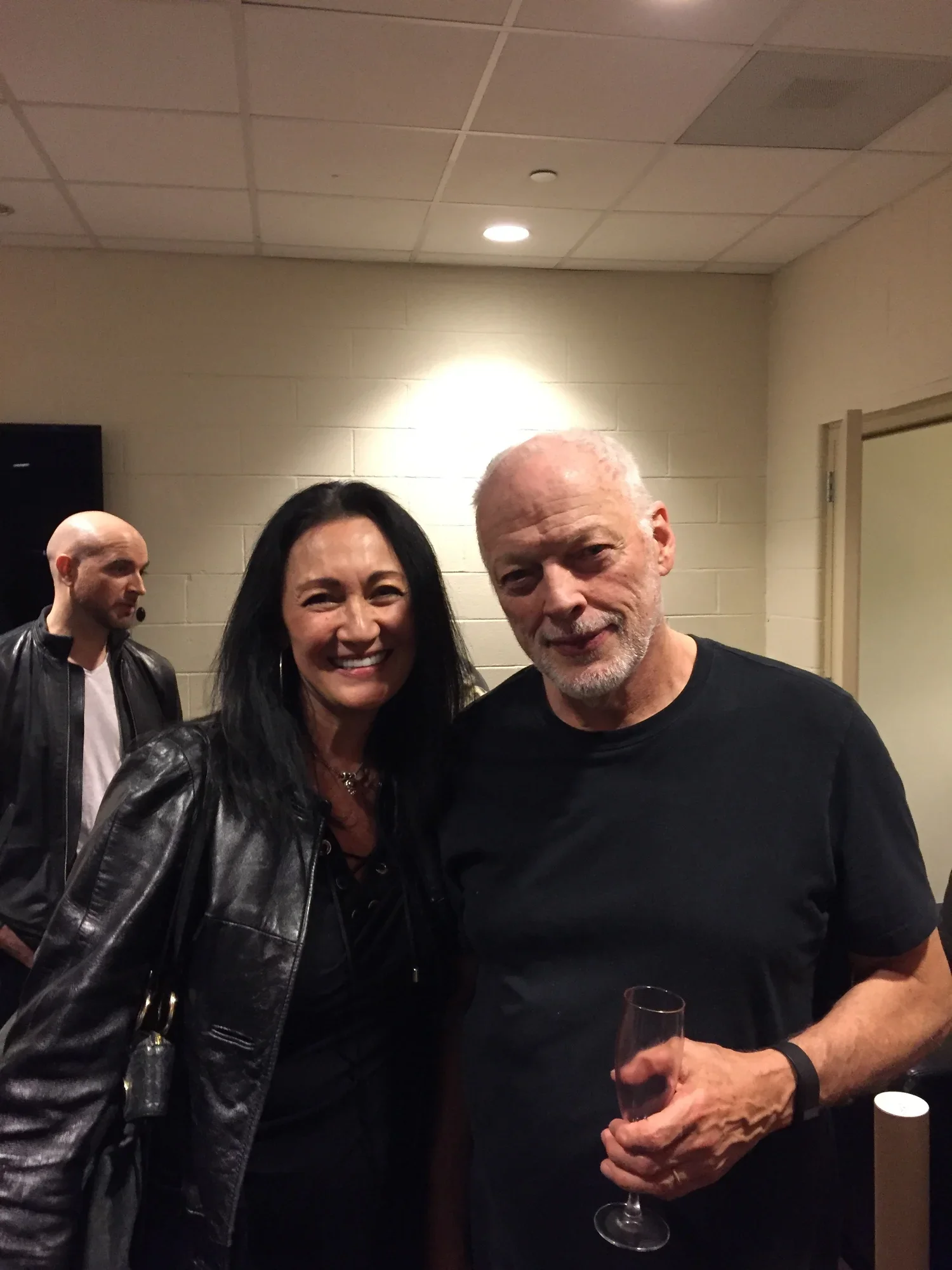 Two people smiling and posing for a photo at an indoor event, with a third person in the background. The woman has long black hair and is wearing a black leather jacket. The man has white hair, a beard, and is also wearing a black shirt. The man in t
