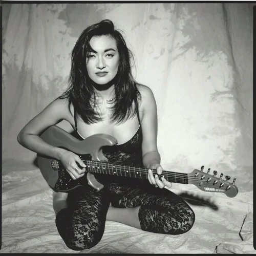 A woman sitting on a bed, holding an electric guitar, wearing lace clothing, with messy hair, in a black-and-white photo.