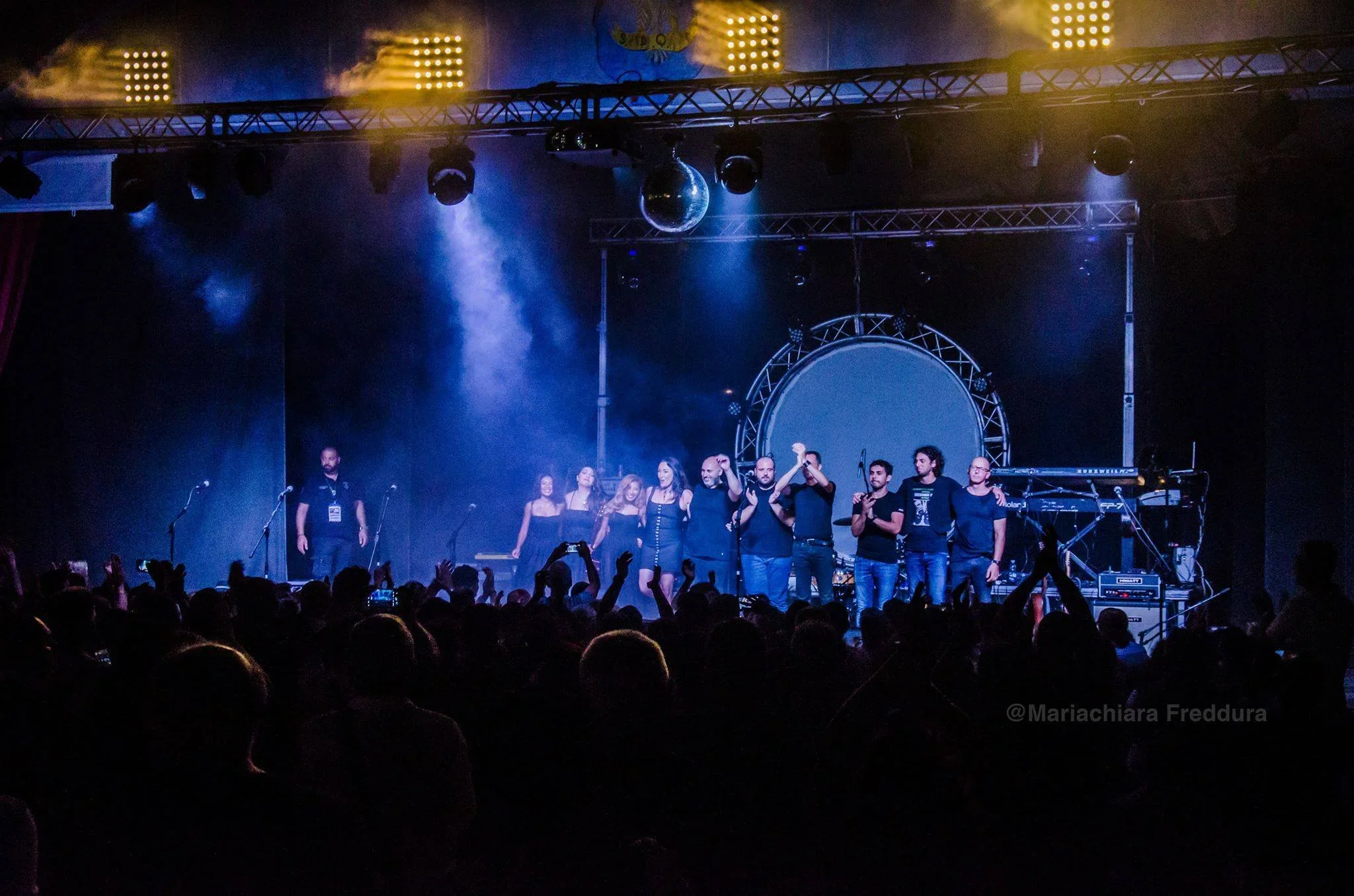 People on stage at a concert or event, with some holding hands and others standing in a row, under colorful stage lighting