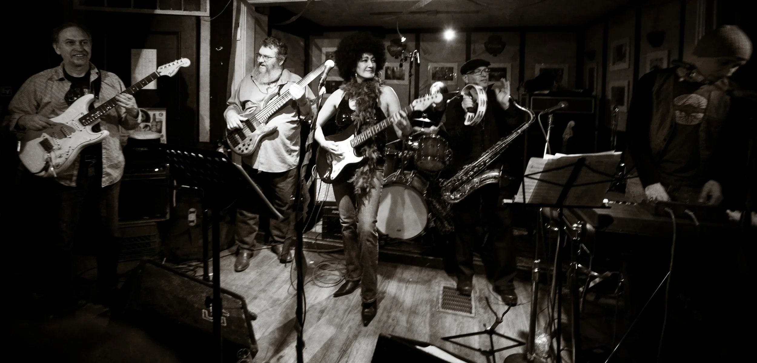 Jazz band performing on stage in a dimly lit venue, featuring musicians playing electric guitar, bass guitar, drums, tambourine, saxophone, and keyboard.