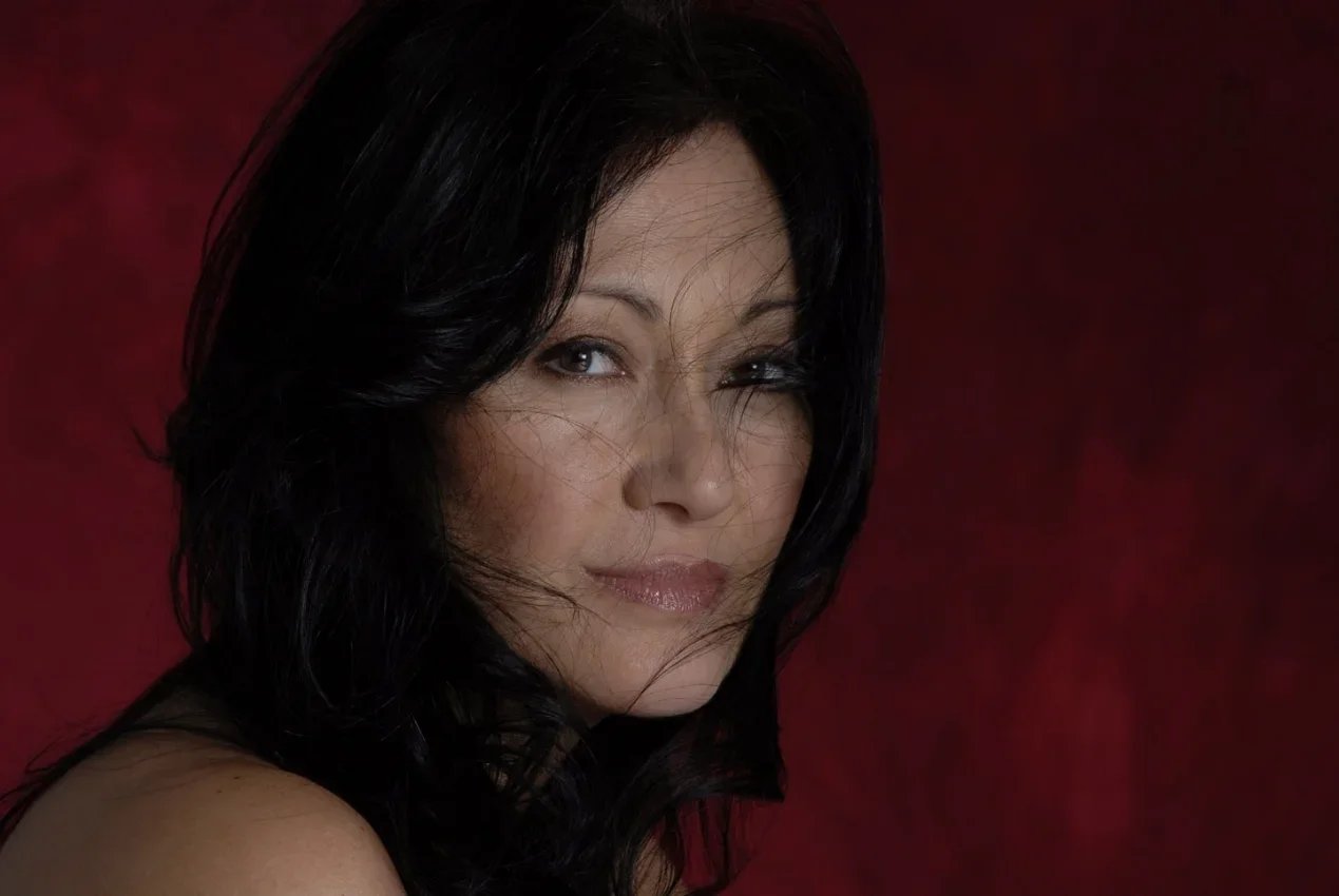 A woman with dark hair and makeup, winking at the camera against a red background.