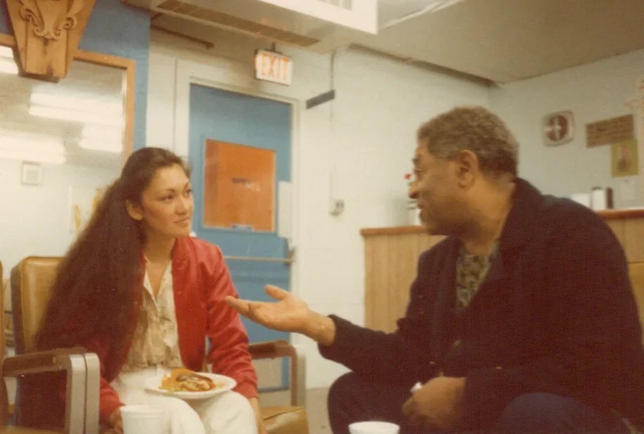 A young woman with long dark hair sitting at a table with a plate of food, listening to an older man with short gray hair and glasses, who is gesturing with his hand while talking in a restaurant or cafe setting.
