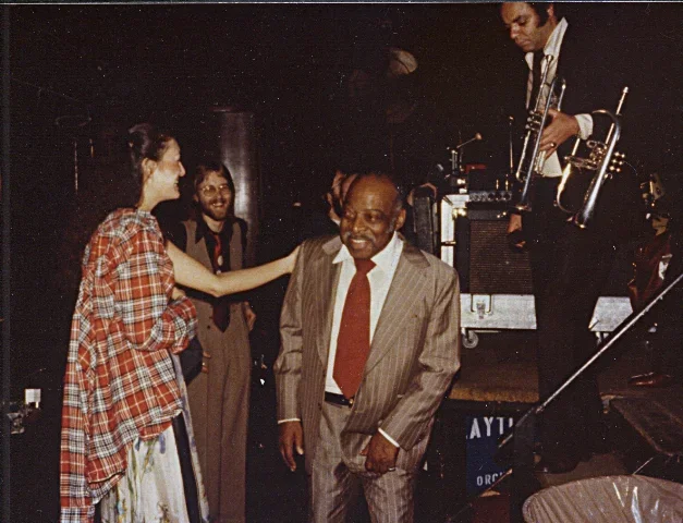 Group of people at a jazz performance, with a woman touching a smiling man in a suit and tie, and a jazz musician playing trumpet on stage.