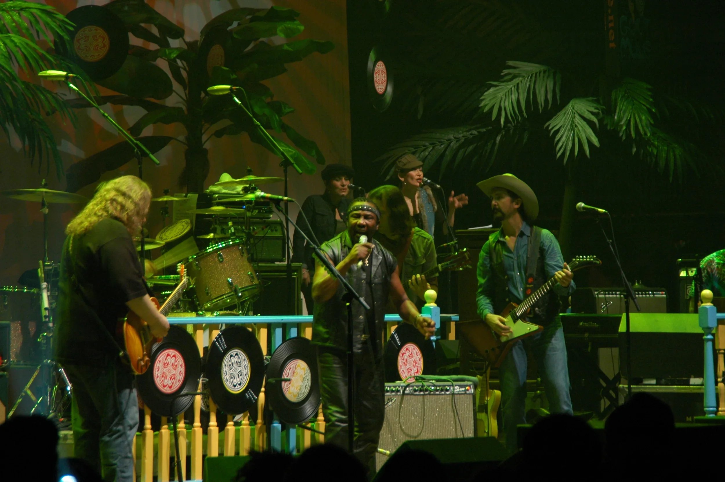 A band performing on stage with various musical instruments, including guitars, drums, and microphones, set against a backdrop of tropical-themed decorations and vinyl records.