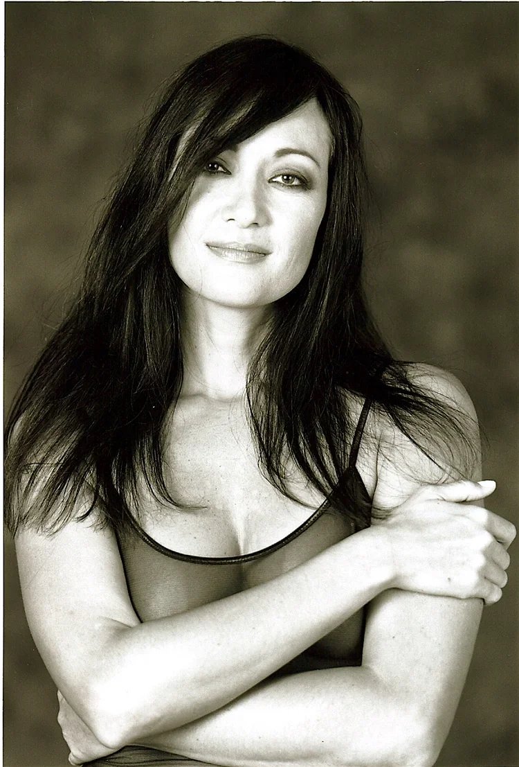 Black and white portrait of a woman with long dark hair, wearing a sleeveless top, looking at the camera with a slight smile, arms crossed.