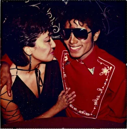 A man and a woman are close together, smiling. The man is wearing large sunglasses and a red shirt with white embroidered designs, and the woman has short dark hair and is wearing earrings and a black outfit.