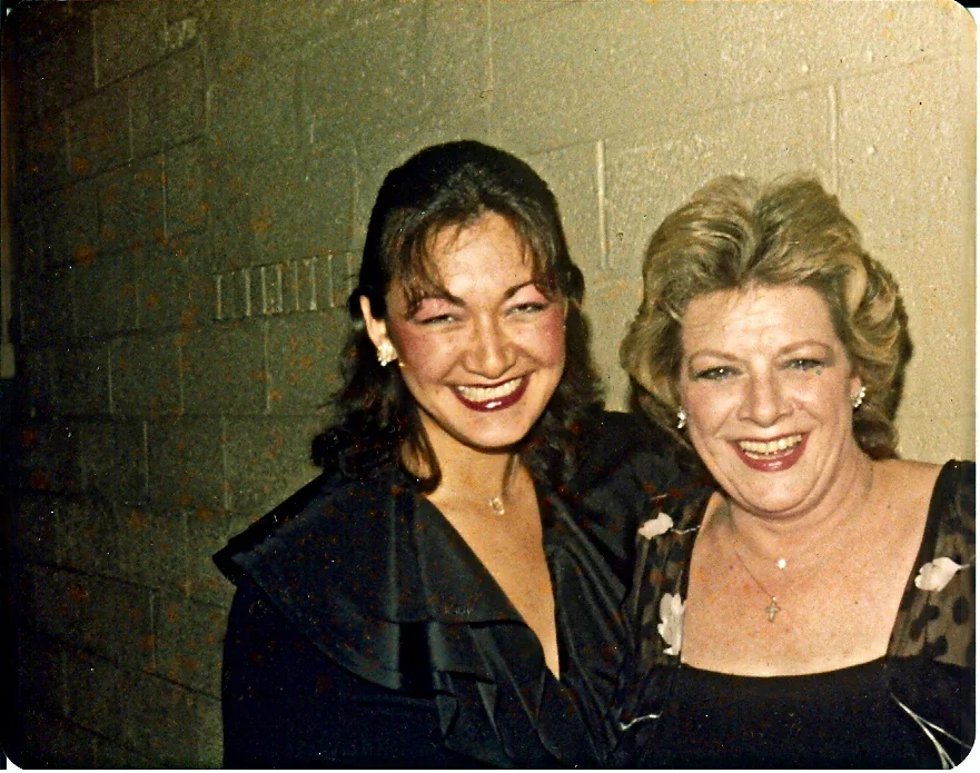Two women smiling and standing together against a plain wall, dressed in dark clothing with jewelry.