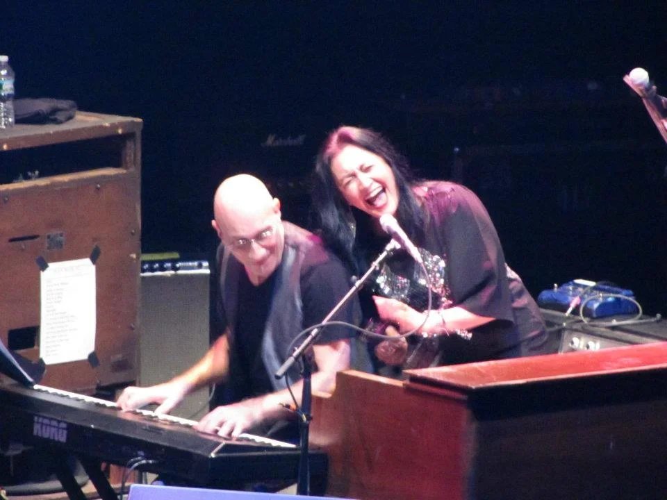 Two musicians, a man playing keyboard and a woman singing into a microphone, performing on stage with a dark background.