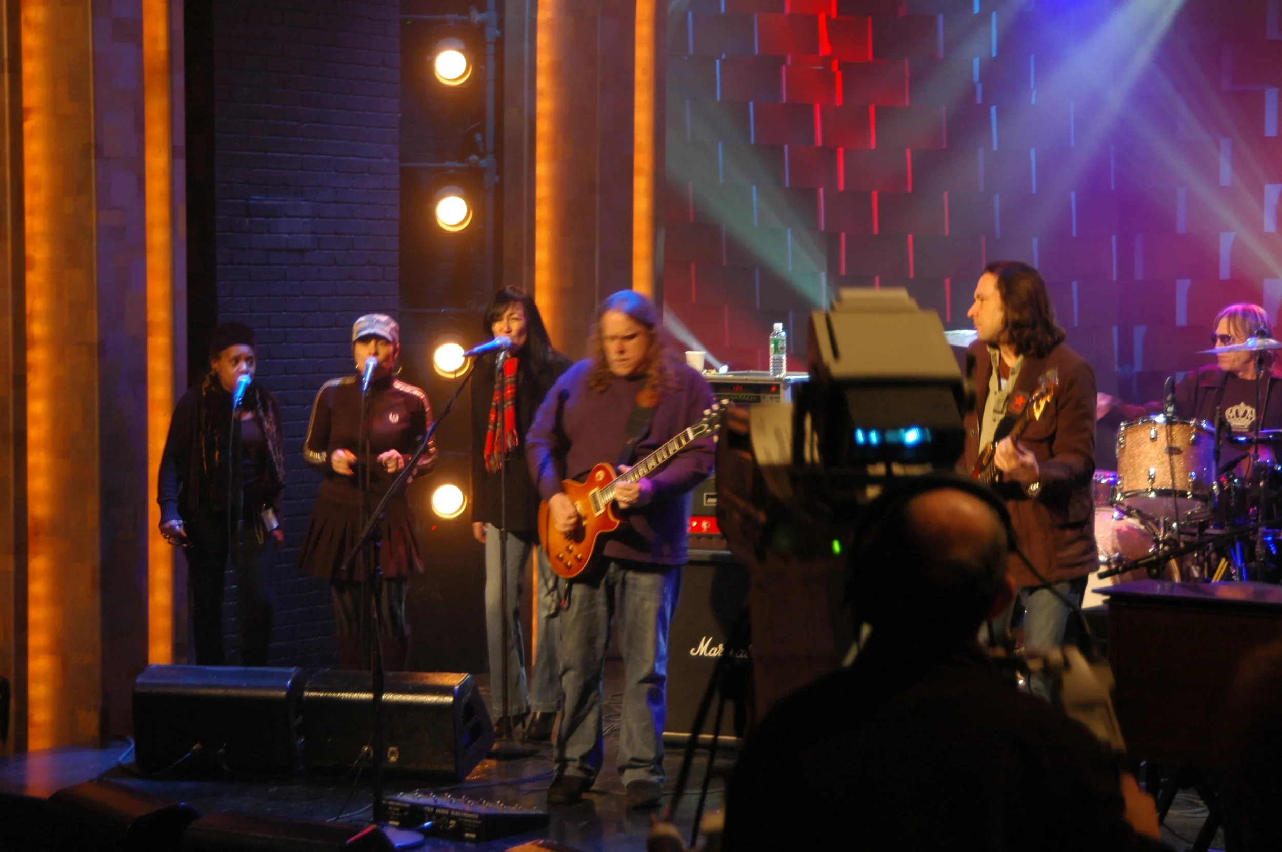 A band performs on stage with five members, including a guitarist in the center, two female vocalists, a male keyboardist, and a drummer in the background. The stage is lit with colorful lights, and there is a dark brick wall with vertical light fixt