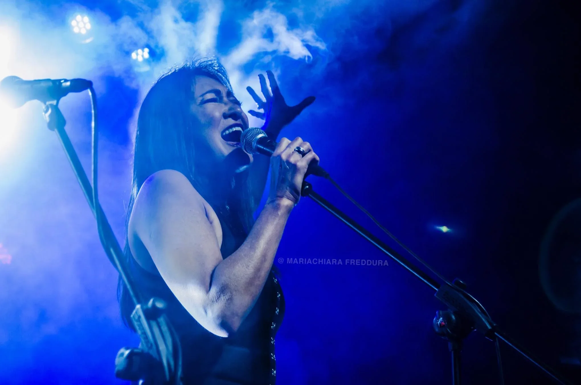 A woman singing passionately into a microphone on stage illuminated by blue stage lighting, with smoke effects around her.