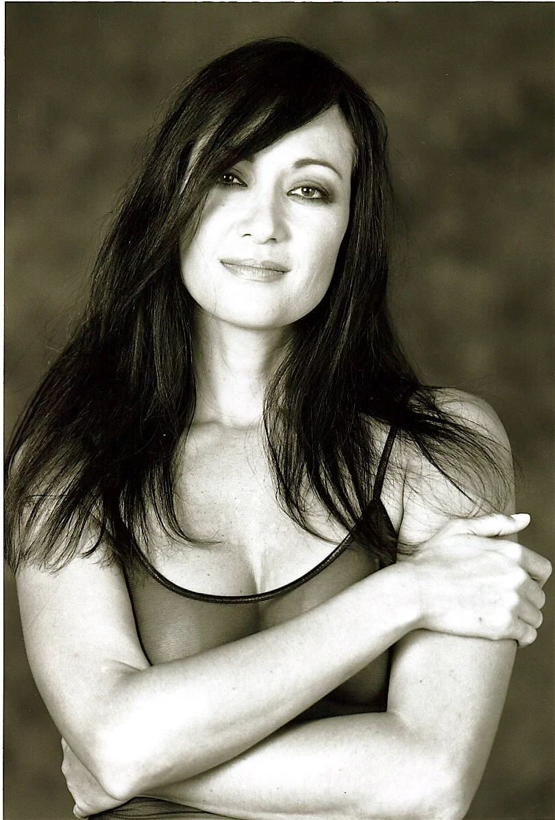 Black and white portrait of a woman with long dark hair, one eye slightly closed, wearing a sleeveless top, with her arms crossed
