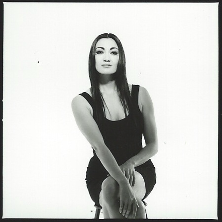 A woman with long dark hair, wearing a sleeveless black top, sitting with legs crossed against a white background.