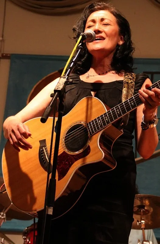 A woman singing and playing an acoustic guitar on stage during a performance.