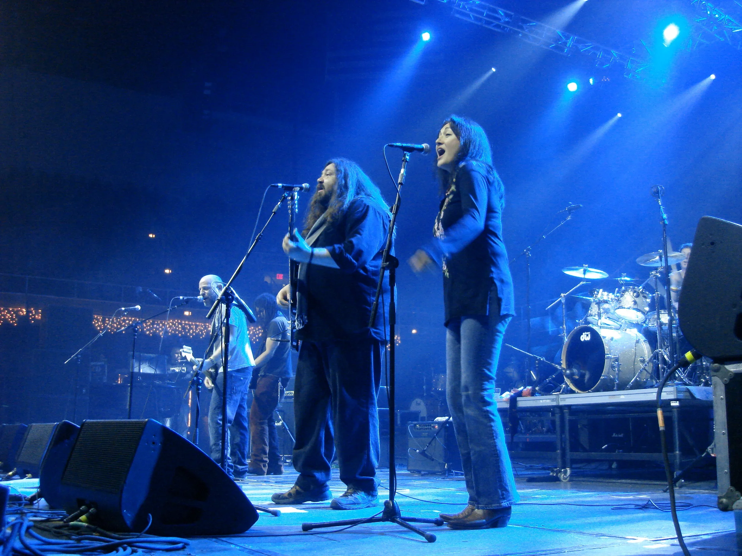 Two women singing into microphones on a stage with blue lighting, with a music band and instruments visible in the background.