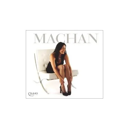 A woman sitting on a white chair wearing a black dress and high heels, smiling, with the word 'MACHAN' written behind her.