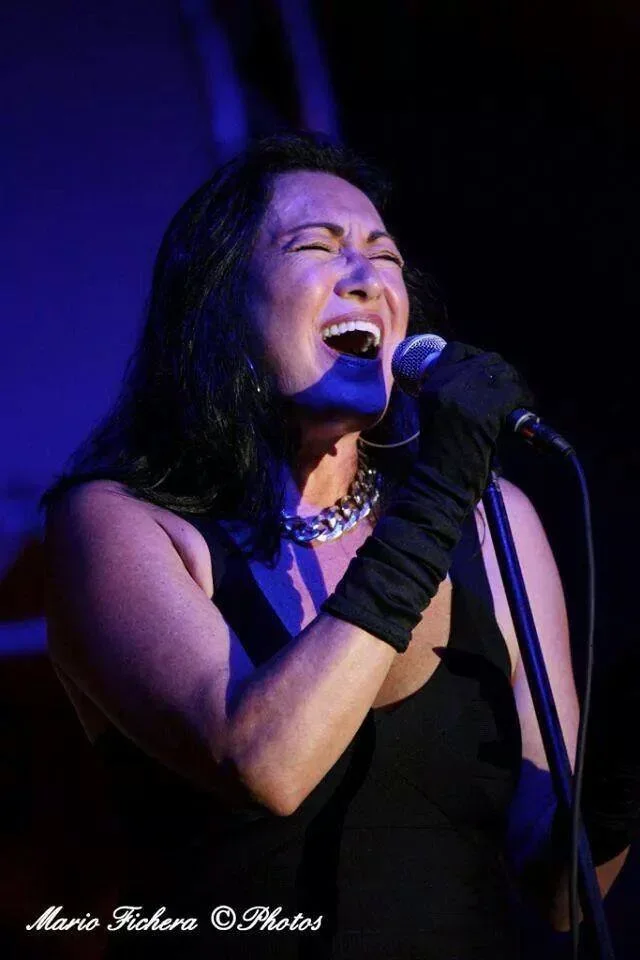 A woman singing passionately into a microphone on stage, wearing black gloves and a black dress, with a necklace and dark hair, illuminated by stage lighting.