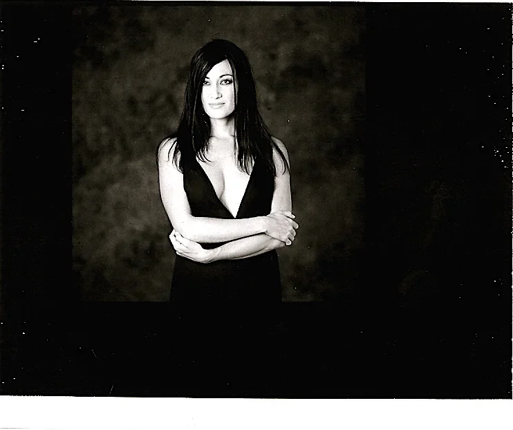 A woman with dark, shoulder-length hair wearing a black dress with a deep V-neck, standing with arms crossed against a dark, textured background.