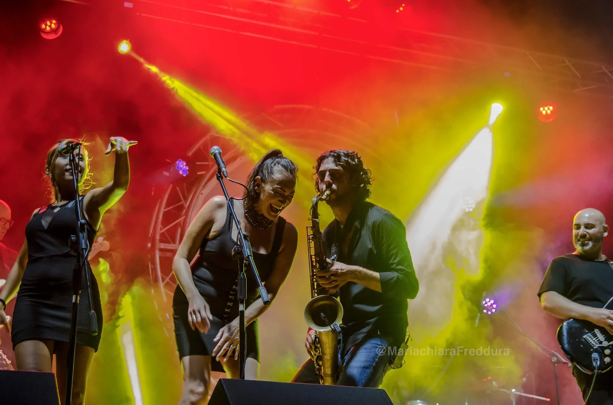 A group of musicians performing on stage, illuminated by colorful stage lights, including yellow and red beams, with some musicians playing instruments and smiling.