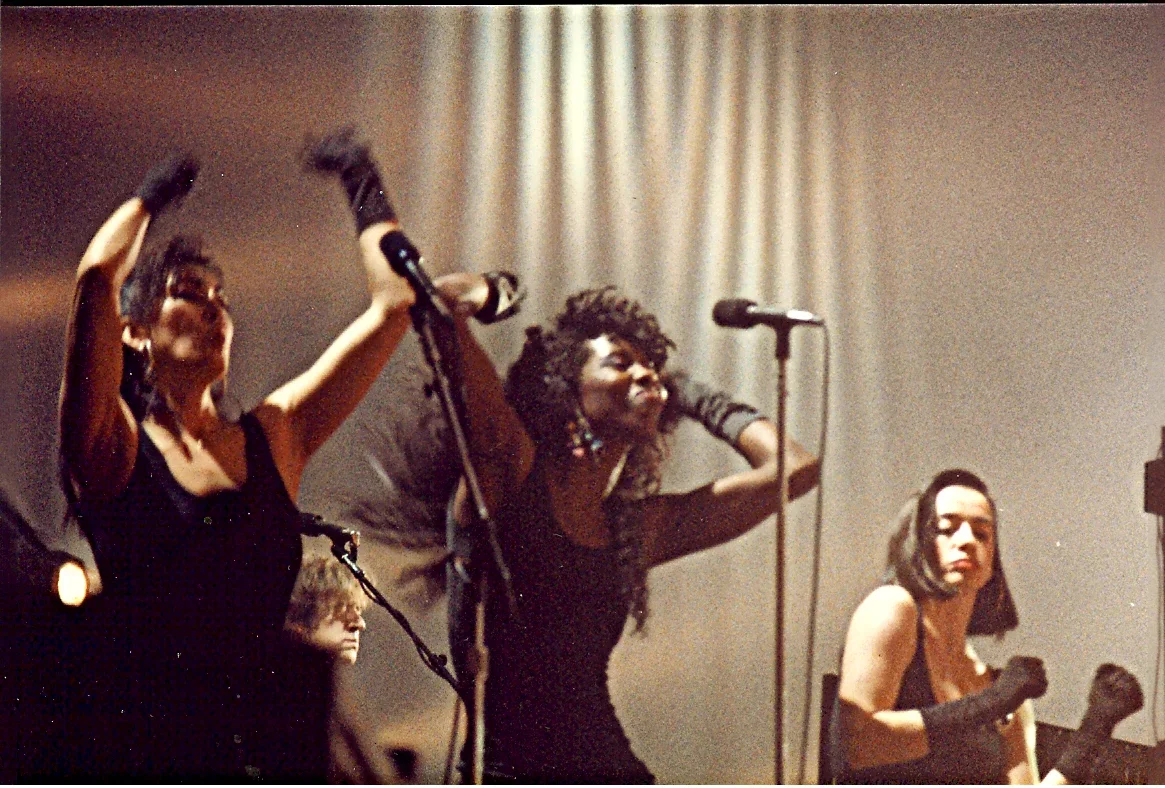 Three women on stage raising their arms in victory or celebration, with a microphone stand in front of them and a white curtain in the background.