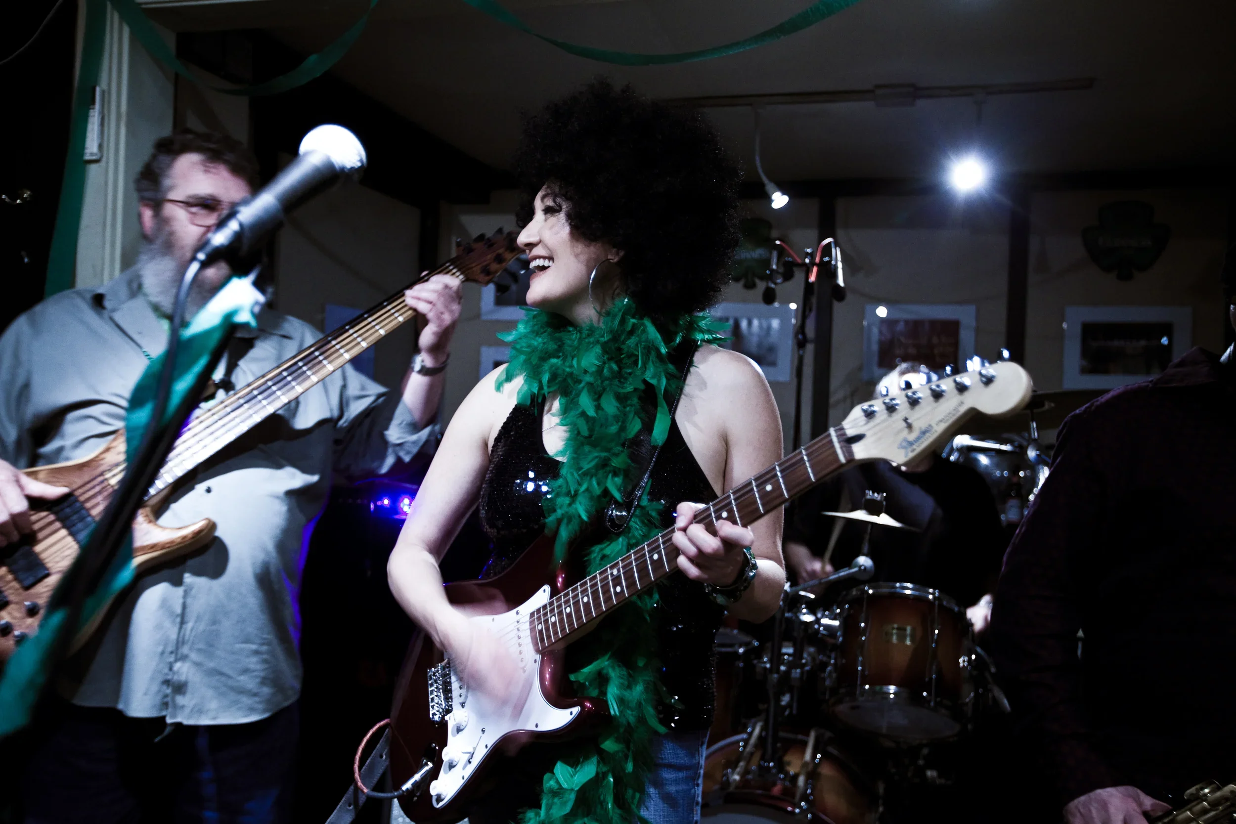 A woman with an afro hairstyle and a green feather boa singing and playing an electric guitar, accompanied by a man with glasses playing an acoustic guitar in a dimly lit indoor setting.