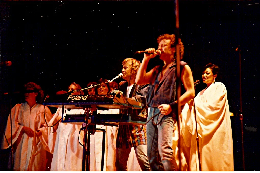 Group of people singing or performing on stage, with some wearing white robes and others in casual clothes, under stage lighting with a dark background.