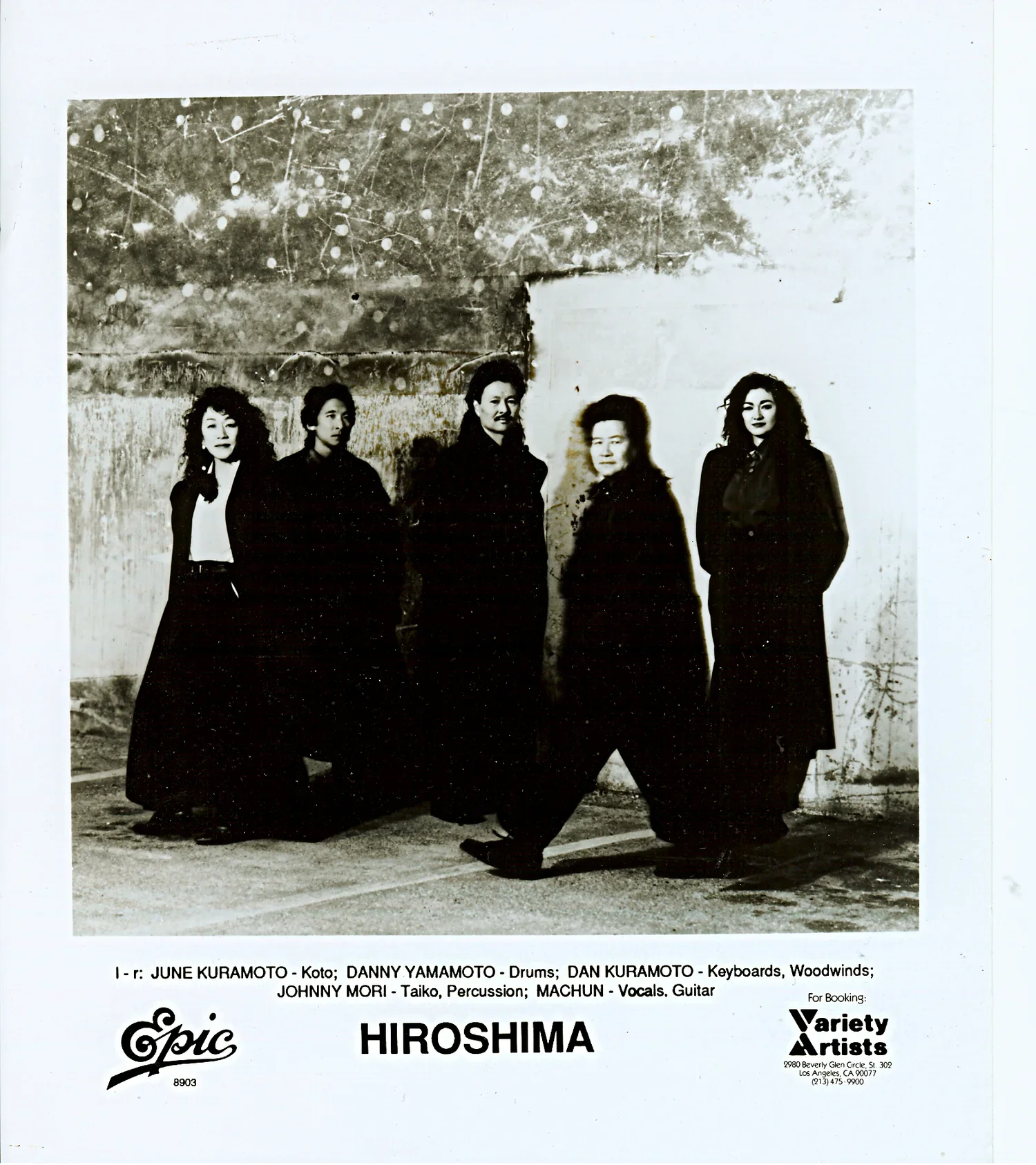 Black and white photograph of five women standing outdoors against a wall with trees and lights above; they are dressed in dark clothing, with some women having curly hair. The text at the bottom indicates the names and roles of the members of the ba