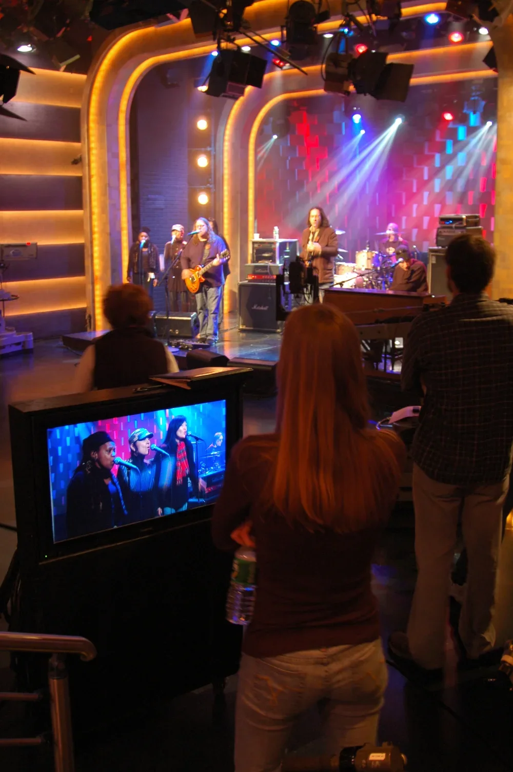 Band performing on stage with audience watching; studio monitor shows singing group, stage lighting with yellow, red, and blue lights.